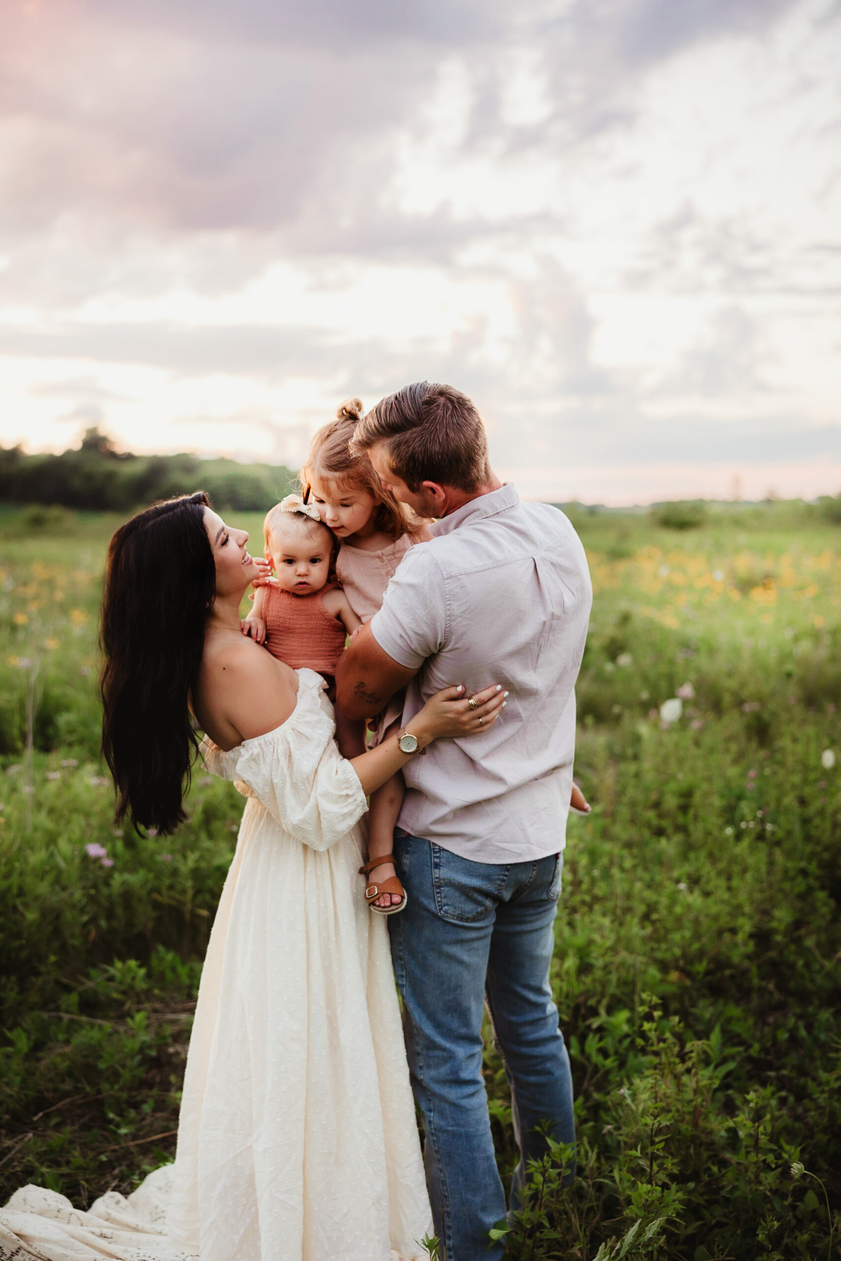 A family of four embraces each other in a wildflower field during sunset.