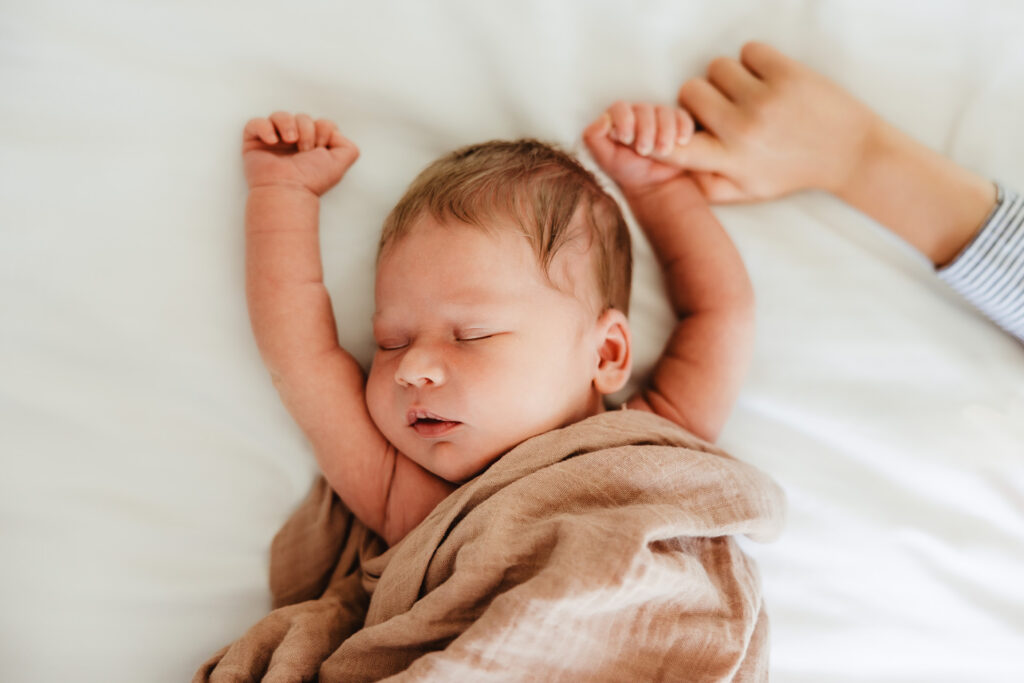 A newborn baby sleeps on the bed while holding his big brothers finger during his Madison Newborn Photography session.